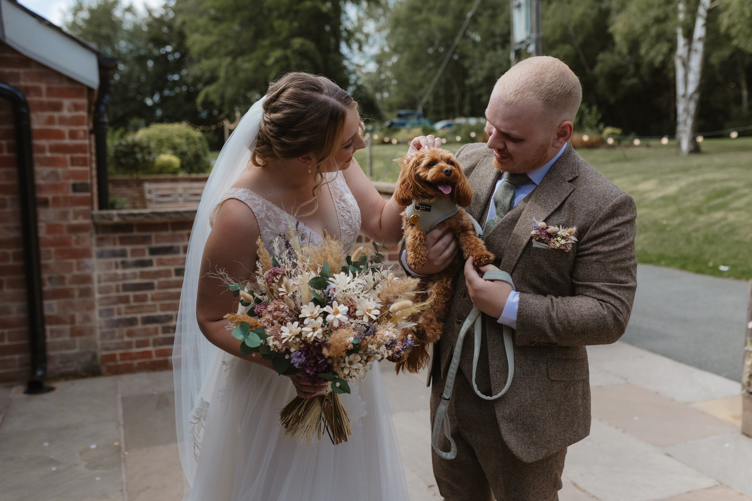 Couple at wedding venue is Staffordshire. Bride is holding a wedding bouquet made and groom is wearing a floral pocket square. Items are made with dried and preserved flowers in pale and pastel colours including sola flowers.